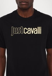 Juodas medvilninis marškinėlis su auksiniu "Justcavalli" logotipu. Dizainas paprastas, turi apvalią iškirptę ir trumpas rankoves. Glotni tekstūra.
