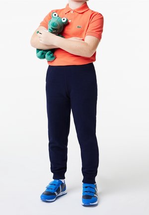 Oranges Polohemd, marineblaue Jogginghose und blaue Sneakers. Hält ein grünes Plüschtier mit großen Augen und offenem Mund.