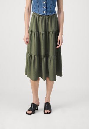 Woman wearing an olive green tiered midi skirt with a drawstring waist, a buttoned blue denim vest, and black open-toe heeled mules.