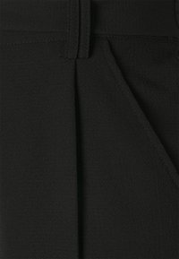 Närbild av svart textil med struktur, med sydda sömmar och en bältesöggla, som visar en del av en ficka på högra sidan.