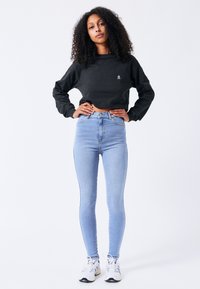 Zwarte cropped sweatshirt met logo, hoge blauwe jeans en witte sneakers met zwarte accenten, tegen een effen achtergrond.