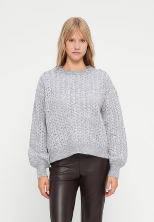 Maglione grigio lavorato a maglia con un motivo testurizzato e maniche a palloncino, con scollatura rotonda e orlo a costine. Indossato sopra pantaloni marroni aderenti.