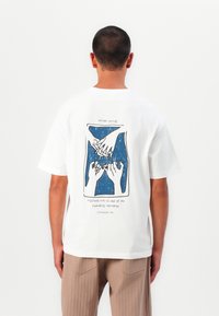T-shirt en coton blanc avec un graphique bleu et noir représentant des mains et du texte au dos. Manches courtes, coupe décontractée et ourlet droit.