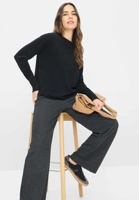 Zwart langemouwen sweater, grijze wijde broek en zwarte sneakers. Beige handtas rustend op de schoot. Houten kruk. Eenvoudige witte achtergrond.