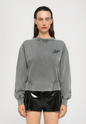 Felpa grigia con polsini e orlo a costine, caratterizzata da un piccolo logo sul petto. Abbinata a pantaloni corti in vinile nero lucido.