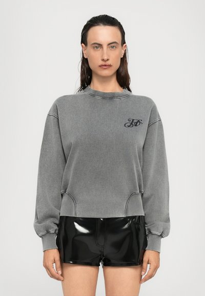 Sudadera gris con puños y dobladillo acanalados, que presenta un pequeño logo en el pecho. Combinada con pantalones cortos de vinilo negro brillante.