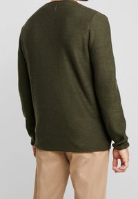 Pull en maille vert foncé avec une surface texturée, col rond, manches longues et ourlet droit, porté sur un pantalon beige.