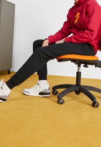 Zwarte joggers, rode hoodie met logo en witte Nike sneakers met grijze accenten. Zittend in een oranje ergonomische stoel op gele vloer.