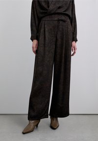 Donkerbruine, geplooide broek met wijde pijpen gemaakt van gestructureerde stof, gecombineerd met puntige bruine laarzen met hak, getoond tegen een lichte achtergrond.