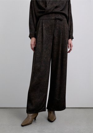 Pantalones anchos de pierna ancha plisados de color marrón oscuro, hechos de tela texturizada, combinados con botas marrones de tacón puntiagudo, exhibidos sobre un fondo claro.