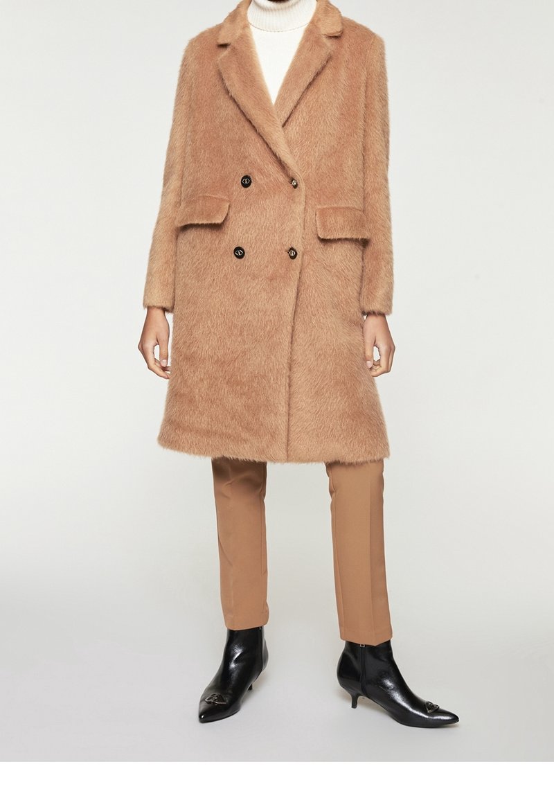 Cappotto doppiopetto in morbido materiale color cammello, con due tasche frontali e bottoni neri, abbinato a pantaloni beige e stivali neri a punta.