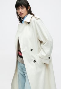 Hvid trenchcoat med sorte knapper, brede reverser og en lige silhuet. Kombineret med en strikket sweater og lyseblå jeans.