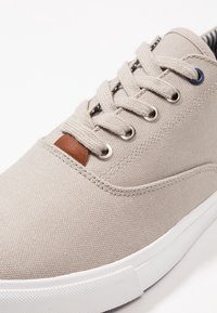 Beige canvas sneakers med vit gummisula, metallöglor, brunt läderdetalj och texturerad yta. Har grå skosnören och inre ränder.