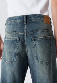 Back view of person wearing faded blue denim jeans and a white ribbed top, highlighting large square back pockets and belt loops.