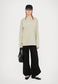 T-shirt de manga longa cinza claro com logótipo, combinado com calças cargo largas pretas e sapatos baixos pretos. Acessório: bolsa preta texturizada e boné.