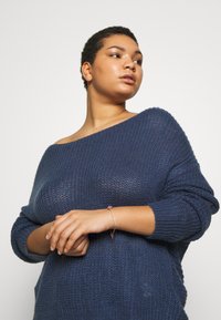 Maglione lavorato a maglia blu navy con design scoperto sulle spalle, vestibilità ampia e tessuto texturizzato. Un delicato braccialetto impreziosisce il polso.