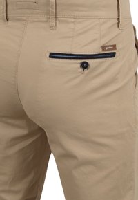 Beige katoenen broek met een achterzak met een donkere knop en een marineblauwe accentstreep. Etiket gevecht in de tailleband. Glad textuur.