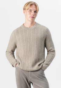 Un pull beige clair avec un design en tricot à câble texturé, doté d'un col rond et de poignets côtelés, associé à un pantalon en velours côtelé gris.