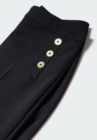 Pantalon noir sur mesure à taille haute avec trois boutons blancs alignés verticalement près de la poche, sur un fond clair.