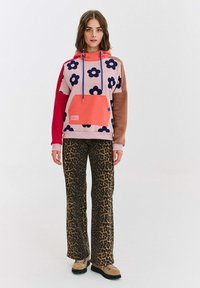 Pestrofarebná mikina s kvetinovým vzorom, červenými a hnedými rukávmi, predná vrecko, v kombinácii s nohavicami s leopardím vzorom. Obuv je svetlohnedá s čiernymi podrážkami.