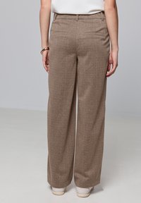 Pantalons amples texturés marron à taille haute, avec deux poches arrière et une coupe décontractée, accompagnés de chaussures de couleur claire.