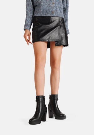 Stivaletti in pelle nera con tacco spesso e fibbia laterale, abbinati a una gonna in ecopelle nera e a un maglione blu lavorato a maglia.