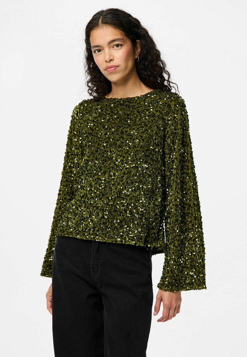 Top lungo a maniche, verde con paillettes, dal fit rilassato e scollatura ampia. Superficie texture con dettagli lucidi e riflettenti. Abbinato a pantaloni neri.