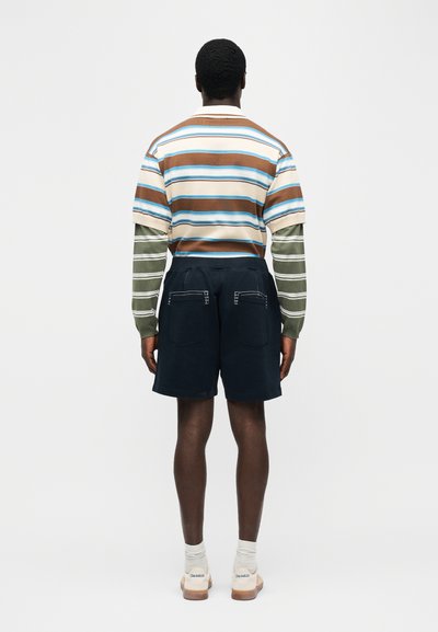 Homme vu de dos portant un polo à manches courtes rayé marron, blanc et bleu sur des manches longues rayées vertes et blanches, un short bleu marine et des chaussures blanches.