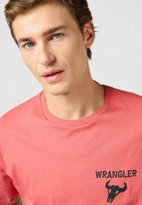Camiseta de algodón color coral con cuello redondo, que presenta un logo negro de "WRANGLER" y un gráfico de calavera en el lado izquierdo del pecho.