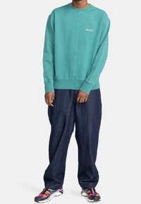 Felpa teal con scollatura rotonda e polsini a coste, abbinata a jeans blu scuro dal taglio morbido e sneakers multicolori.