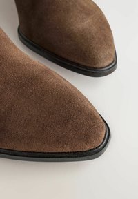 Ruskeat mokkasiinikengät, joissa on musta reuna pohjassa, kuvattuna läheltä kulma-alirosta vaalealla taustalla.