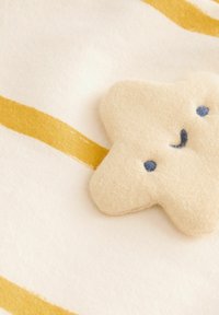 Parche en forma de estrella de felpa beige con detalles de costura en azul, colocado sobre una suave tela color crema con dos franjas doradas.