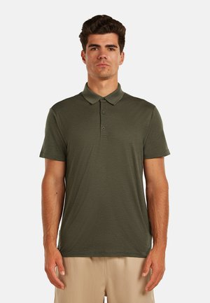 Mann steht und trägt ein olivgrünes kurzärmeliges Poloshirt und beige Hose vor einem einfarbigen hellen Hintergrund.