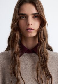 Brauner gestrickter Pullover mit rundem Ausschnitt, über einem burgunderfarbenen Hemd mit Kragen getragen. Welliges Haar und natürliches Makeup betonen den Look.