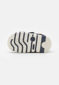 Suola di scarpa in gomma, prevalentemente bianca con accenti blu navy. Presenta scanalature ondulate, fori di ventilazione e marchi del brand.