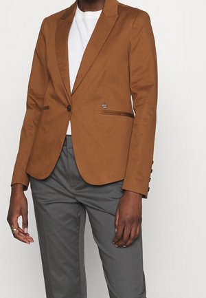 Person iført en brun enkeltknappet blazer over en hvid skjorte kombineret med grå bukser, stående mod en ensartet baggrund.