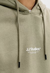 Felpa con cappuccio verde oliva con coulisse, texture morbida, caratterizzata da un logo bianco "JJ Studios" e dettagli neri alle estremità della coulisse.