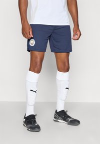 Shorts de foot bleu marine avec le logo de Manchester City, associés à des chaussettes blanches jusqu'aux genoux et des chaussures de foot noires avec un design texturé.