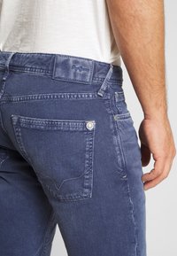 Tmavě modré džíny z denimu se mírně vybledlým povrchem. Obsahují prošívané zadní kapsy a koženou nášivku s logem na pásku.