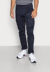 Granatowe spodnie cargo z wieloma kieszeniami, o dopasowanym kroju i trwałym materiale. Połączone z białymi butami sportowymi, z czarnymi akcentami i czerwonym logo.