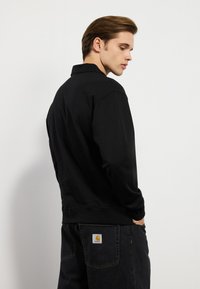 Svart hettegenser med klassisk krage, laget av et mykt stoff. Har lange ermer og ribbestrikket kant. Stylert med mørke jeans.