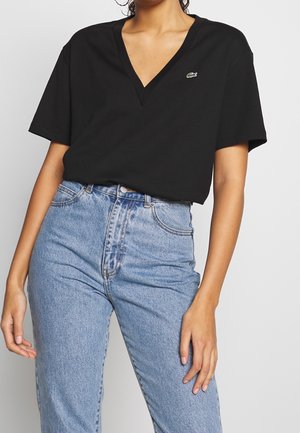 Kobieta ubrana w czarny krótki t-shirt z dekoltem w serek, z małym zielonym logo krokodyla oraz jasnoniebieskie, wysokie spodnie jeansowe.