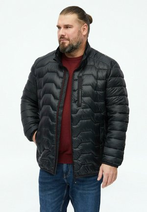 Mann mit Bart und zurückgebundenen Haaren, der eine schwarze gesteppte Jacke über einem roten Pullover und blauen Jeans trägt, steht mit den Händen in den Taschen.