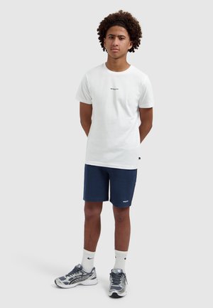 Jonge man staat met de handen achter de rug, draagt een wit T-shirt, marineblauwe korte broek, witte sokken en grijze hardloopschoenen op een effen achtergrond.