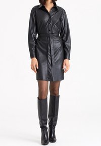 Vestito camicia in finta pelle nera con maniche lunghe, chiusura a bottoni davanti, vita con cintura e stivali neri fino al ginocchio. Texture liscia, vestibilità su misura.