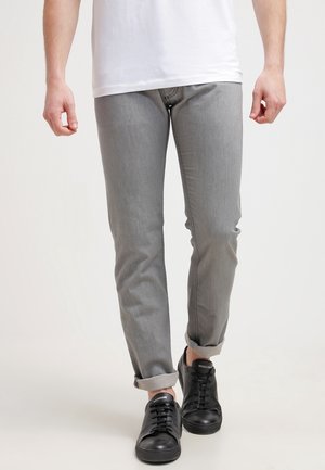 Man draagt lichtgrijze opgerolde spijkerbroek, een wit T-shirt en zwarte sneakers, lopend op een effen lichte achtergrond.