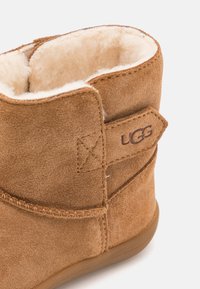 Brune semsket ankelstøvler med en blød kremfarget foring, som har en sydd søm og UGG-logo på siden. Sålen er laget av lysebrunt gummi.