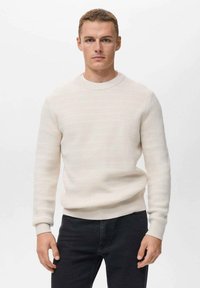 Suéter de crema texturizado con un cuello redondo acanalado y puños, combinado con jeans negros. La tela tiene un sutil patrón tejido.