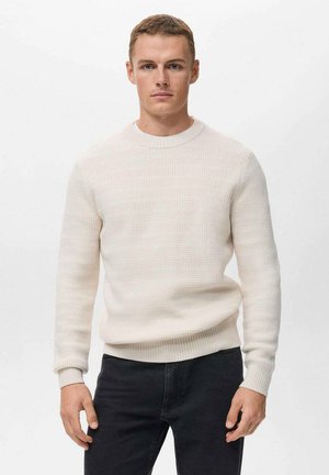 Suéter de crema texturizado con un cuello redondo acanalado y puños, combinado con jeans negros. La tela tiene un sutil patrón tejido.