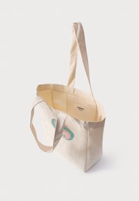 Beige lerret tote veske med to solide håndtak, med et fargerikt rundt logodesign på den ene siden og en åpen topp for enkel tilgang.